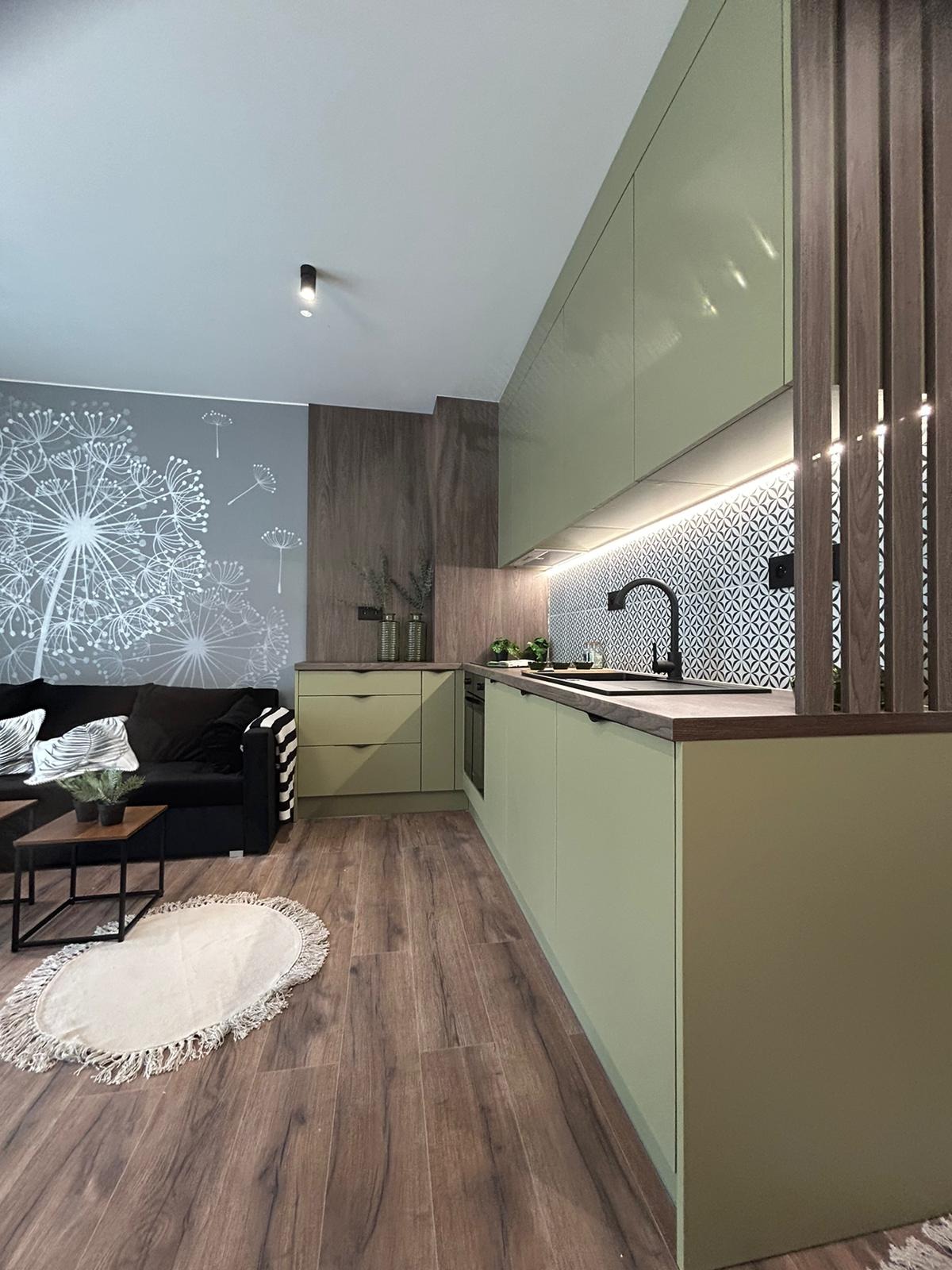 Nowoczesna kuchnia w odcieniach zieleni z drewnianymi akcentami, wzorzyste płytki nad blatem, parkiet i salon z sofą w tle. Całość tworzy spójną, stylową przestrzeń.