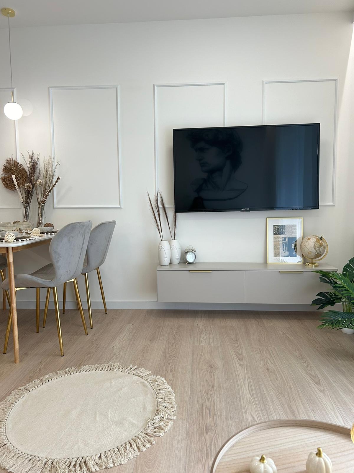 Jasne wnętrze salonu z drewnianą podłogą, szarymi krzesłami z złotymi nogami i telewizorem z wizerunkiem rzeźby Dawida na ścianie. Minimalistyczny design.