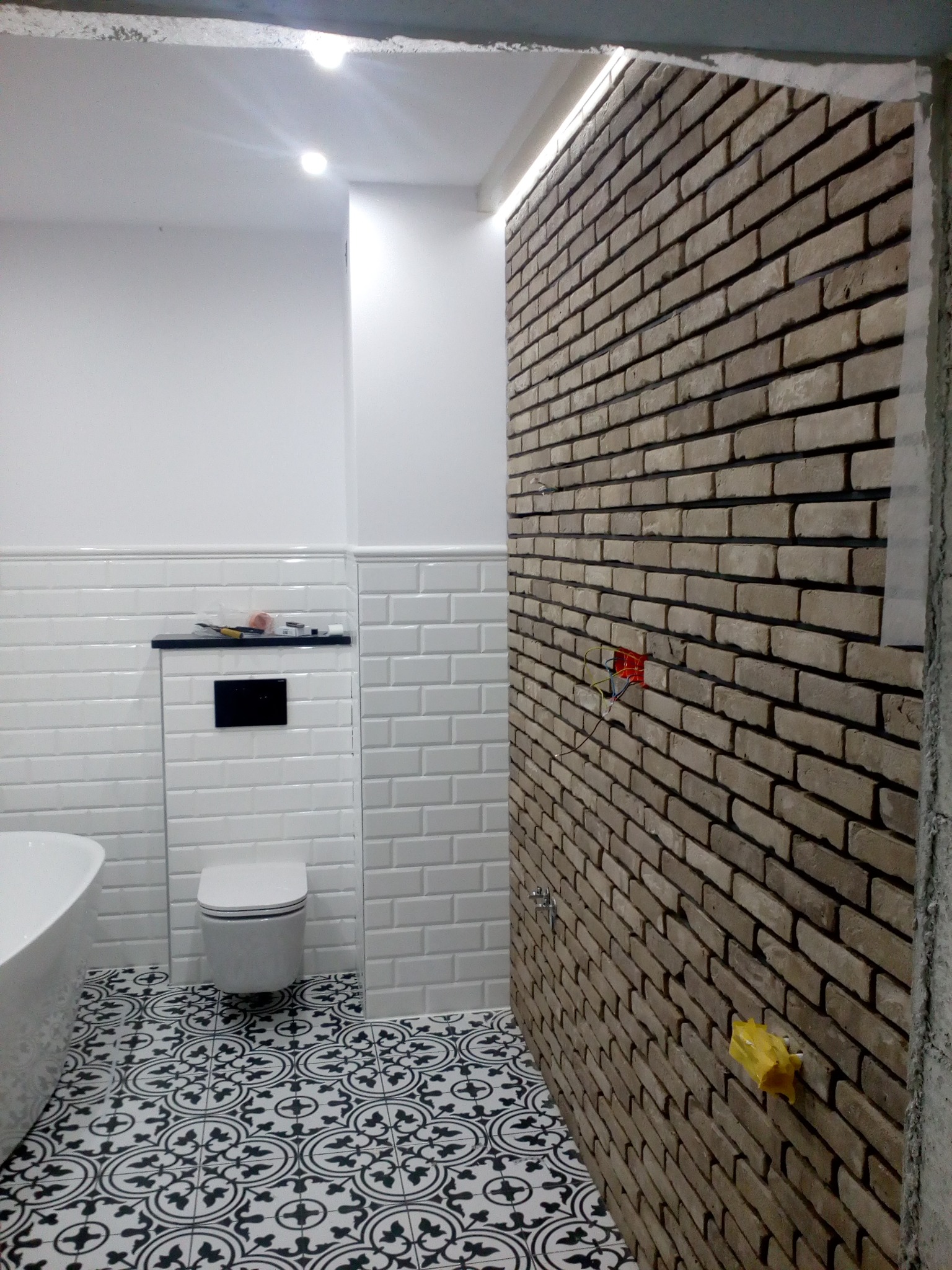 Wnętrze łazienki w trakcie wykończenia: ściana z cegieł klinkierowych z widoczną instalacją elektryczną, białe płytki typu metro, czarno-biała wzorzysta podłoga, częściowo widoczna wanna i toaleta.