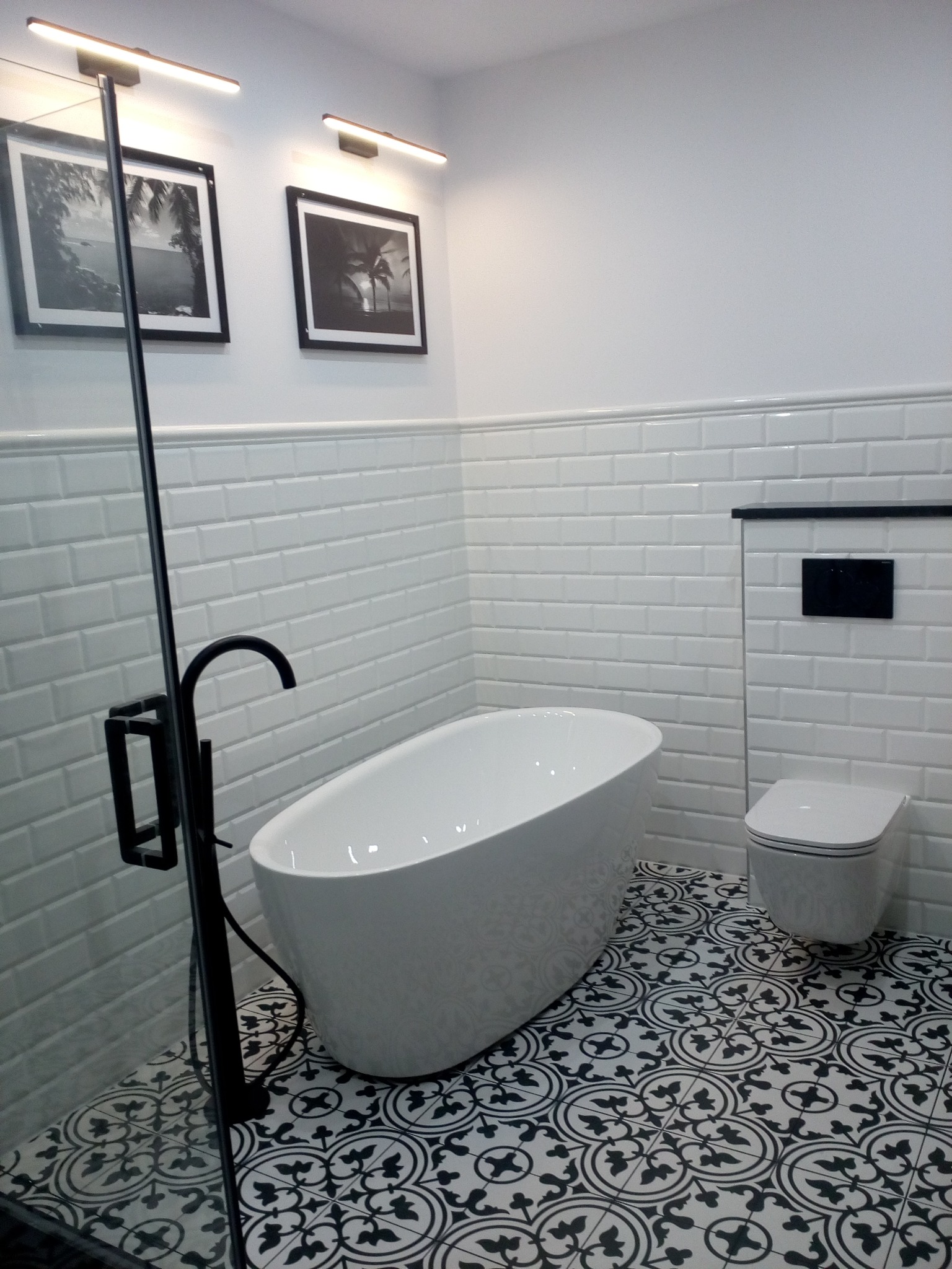 Biała wolnostojąca wanna i podwieszana toaleta w nowoczesnej łazience z białymi, prostokątnymi płytkami ściennymi i czarno-białą wzorzystą podłogą; nad wanną czarna bateria podłogowa, a na ścianie...