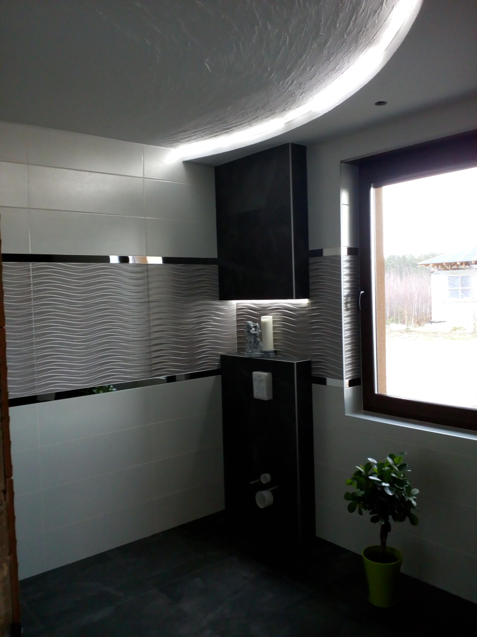 Nowoczesna łazienka z białymi i szarymi płytkami, dekoracyjnym paskiem z motywem fali, podwieszaną toaletą w czarnej obudowie, oświetleniem LED na suficie i rośliną doniczkową przy oknie.