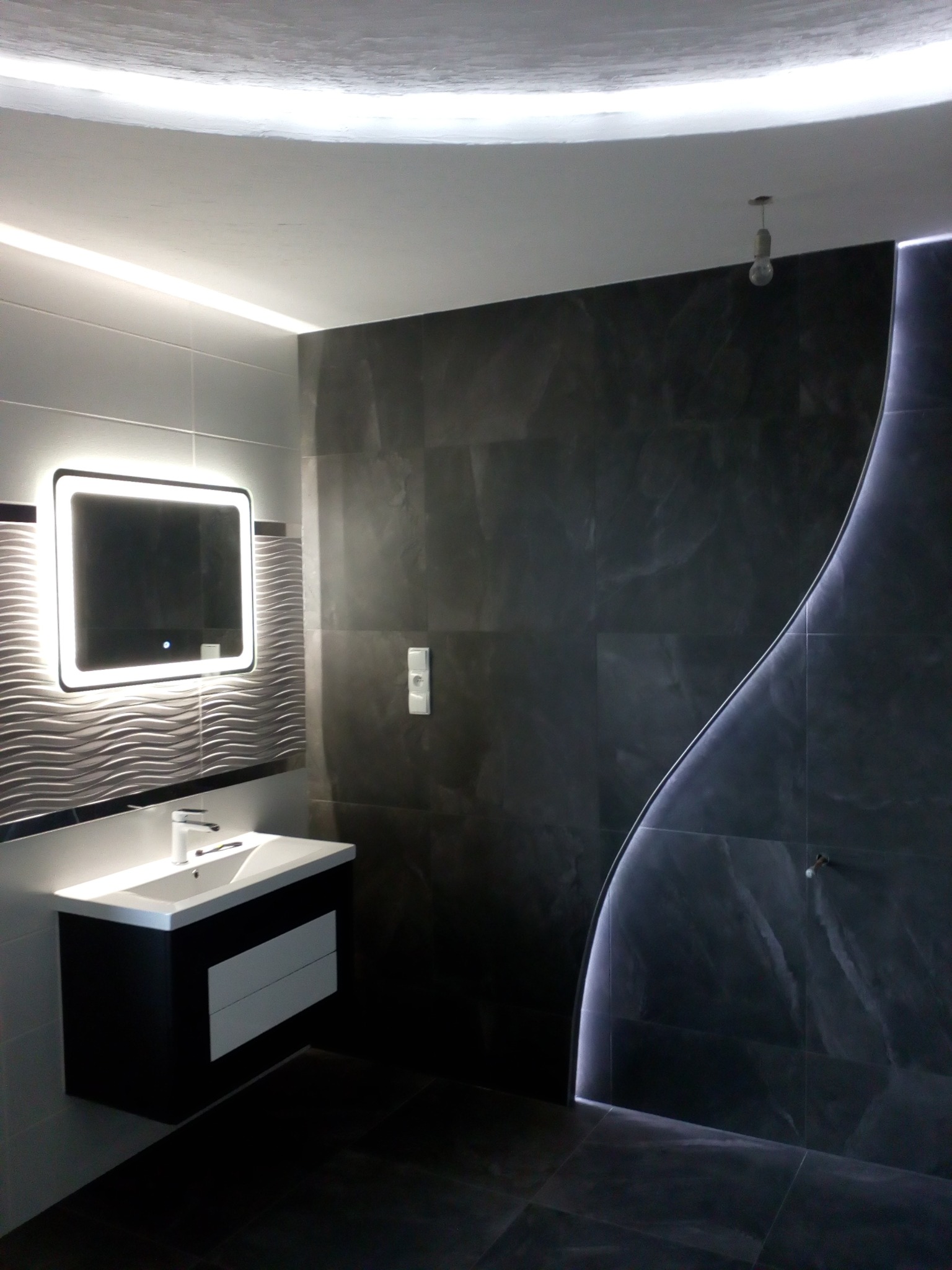 Nowoczesna łazienka z czarno-białymi płytkami, podświetlanym lustrem i dekoracyjnym oświetleniem LED wzdłuż ściany prysznicowej, oraz wisząca umywalka z szafką.