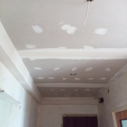 Firma remontowa - Sufit z płyt kartonowo-gipsowych, z widocznymi miejscami łączeń wypełnionymi gładzią szpachlową, otwór na oświetlenie z wystającym przewodem, widoczne niedoskonałości na ścianach i brakujące listwy...