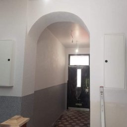 Firma remontowa - Korytarz w trakcie prac w Skarżysko-Kamiennej: świeżo pomalowane ściany na biało z szarym pasem na dole, widoczny łuk, zabezpieczone drzwi wejściowe folią i poziomica oparta o ścianę.