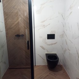 Firma remontowa - Nowoczesna łazienka z kabiną prysznicową z czarną ramą, toaletą w kolorze czarnym i płytkami imitującymi marmur na ścianach oraz drewnopodobną podłogą ułożoną w jodełkę.