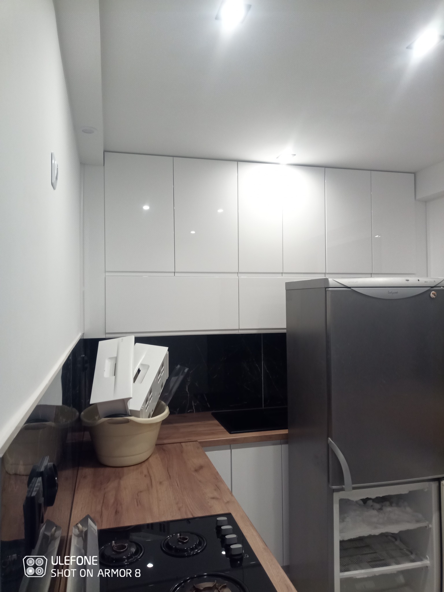 Nowoczesna, jasna kuchnia z białymi, błyszczącymi szafkami, drewnianym blatem, czarnym panelem nad blatem i szarą lodówką. Na blacie stoi wiadro z białymi elementami, prawdopodobnie fragmentami...