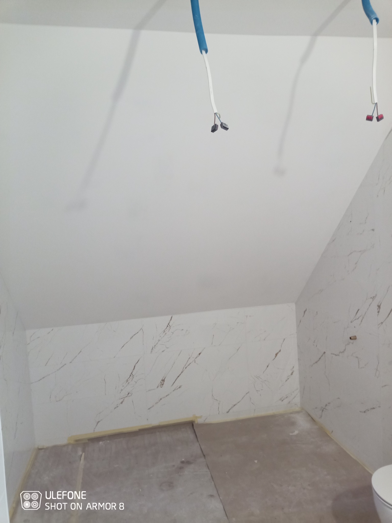 Wnętrze łazienki w trakcie prac wykończeniowych, ściany wyłożone płytkami imitującymi marmur, widoczne wystające przewody elektryczne i przygotowane podłoże pod posadzkę.