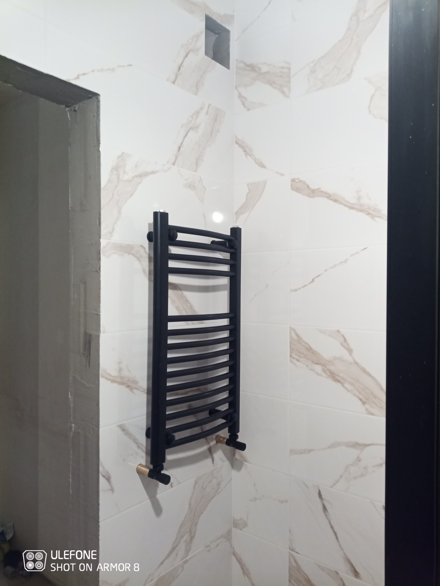 Czarny, drabinkowy grzejnik łazienkowy zamontowany na ścianie wyłożonej białymi płytkami imitującymi marmur, widoczny fragment surowego wykończenia ściany obok futryny drzwi.