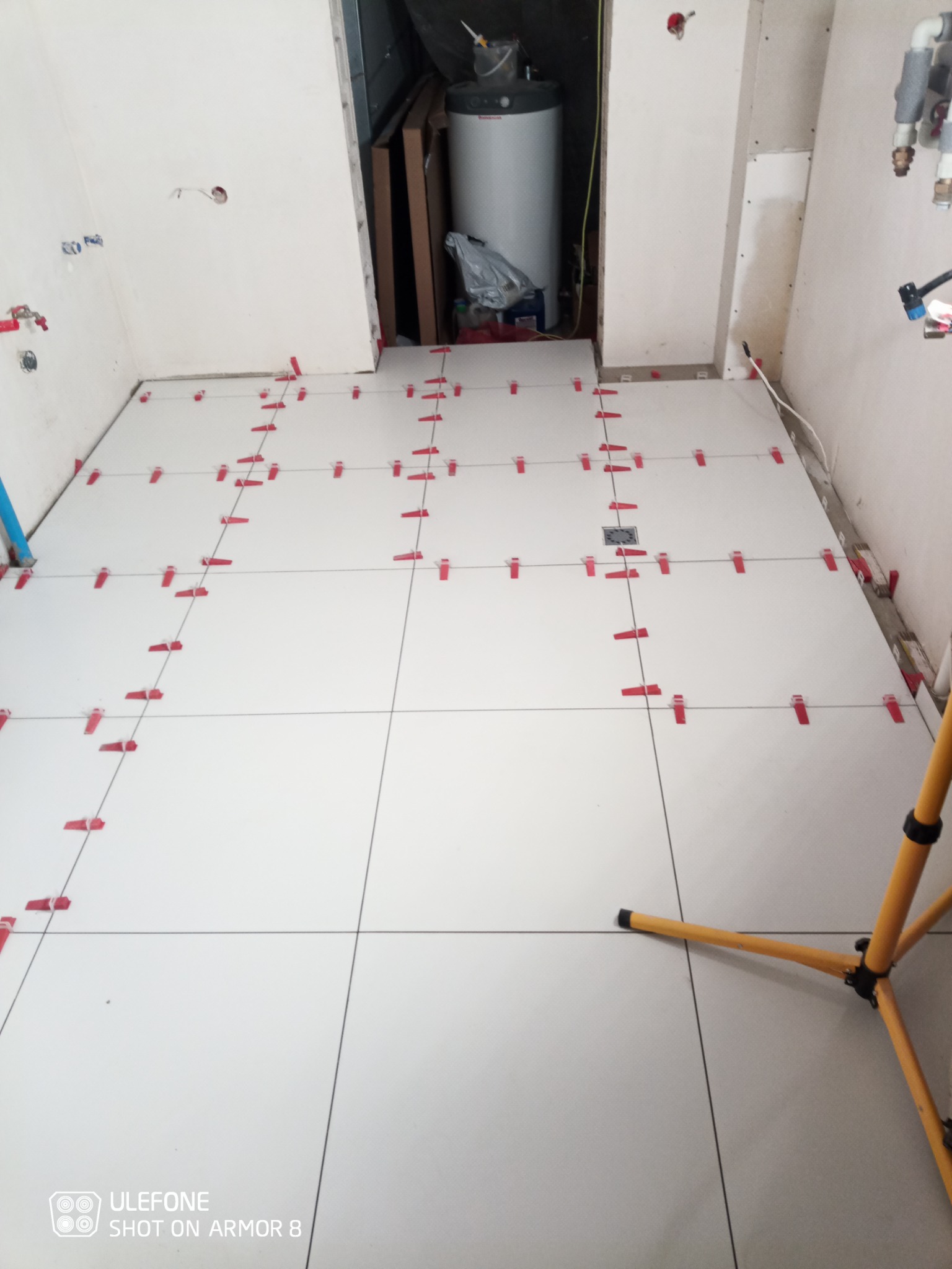 Białe płytki podłogowe ułożone w rzędach, z czerwonymi klipsami poziomującymi, widoczne rury i bojler w tle, częściowo wykończone ściany, lampa robocza z żółtym statywem w prawym dolnym rogu.