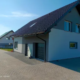 Nowoczesny dom jednorodzinny z ciemną dachówką i szarą elewacją, widok na podjazd wyłożony szarą kostką brukową, błękitne niebo w tle.