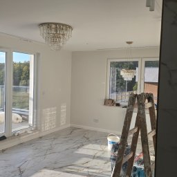 RADOSŁAW LANCZEWSKI - Jasne wnętrze w trakcie wykańczania: marmurowa podłoga, białe ściany, duże okna balkonowe z widokiem na zieleń, na balkonie leżący biały pies, drabina malarska i akcesoria malarskie w rogu pokoju...