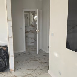 RADOSŁAW LANCZEWSKI - Wykończone wnętrze z marmurową podłogą i widokiem na łazienkę z okrągłym lustrem, białe ściany, czarny worek na śmieci w rogu pomieszczenia.