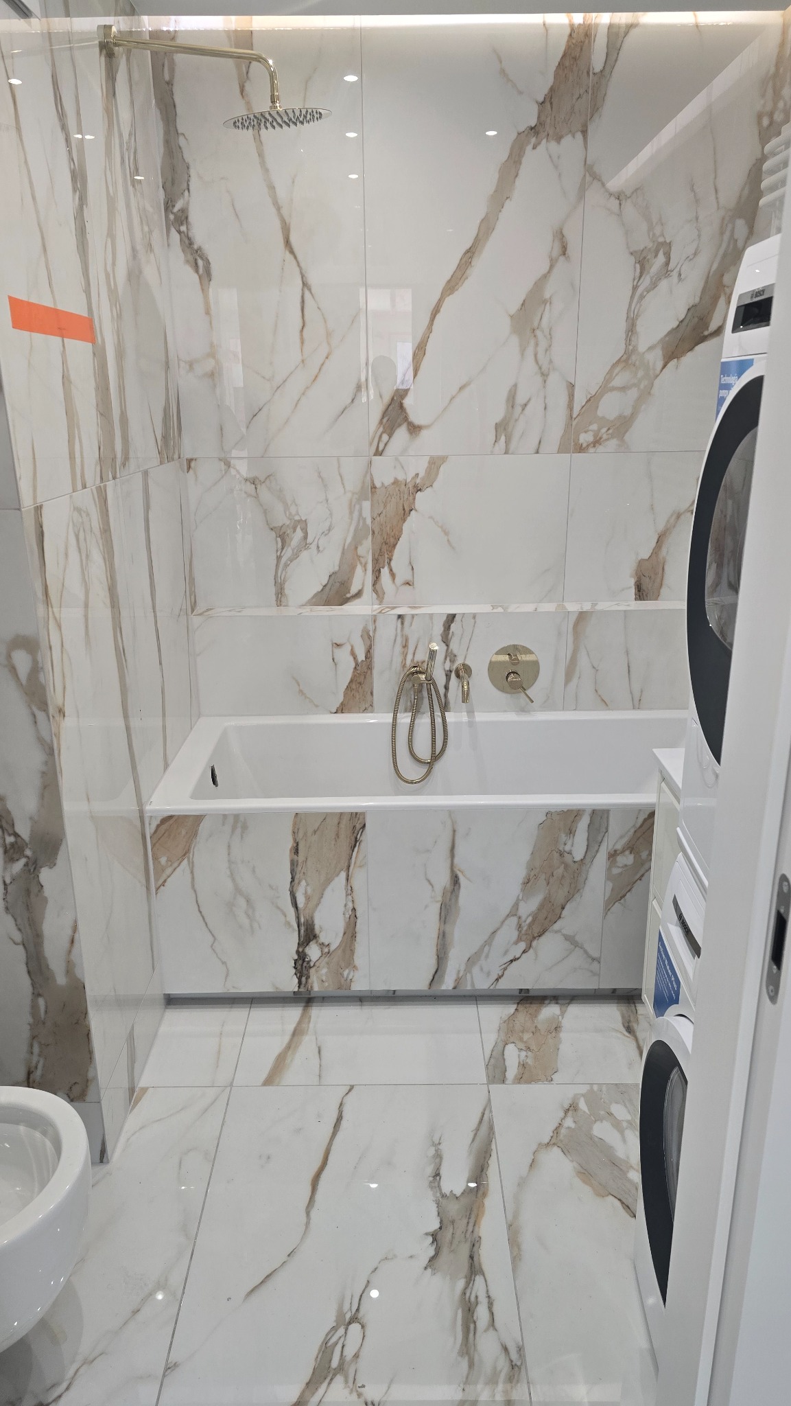 Nowoczesna łazienka z wanną obłożoną płytkami imitującymi marmur, deszczownicą i złotą armaturą, widok na pralkę z boku.