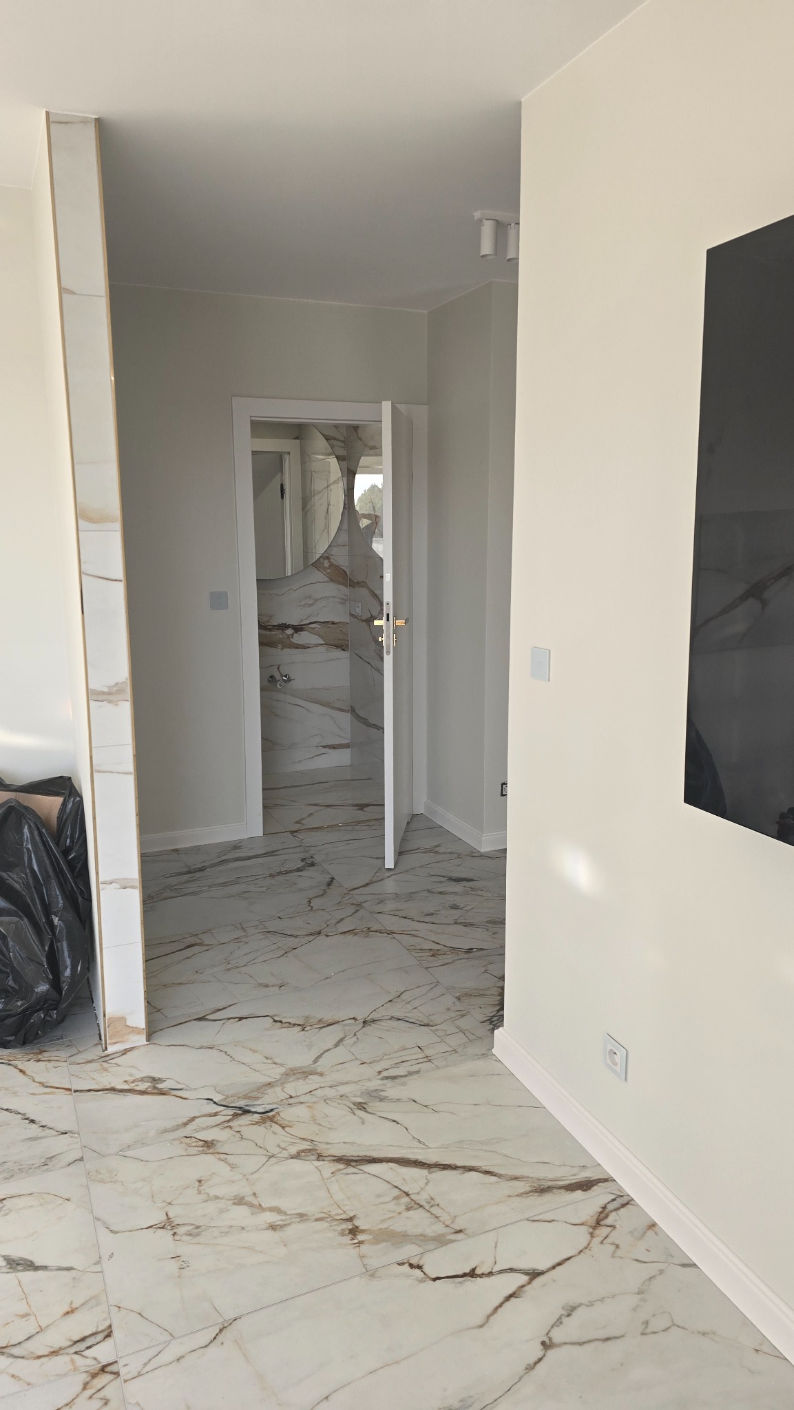 Wykończone wnętrze z marmurową podłogą i widokiem na łazienkę z okrągłym lustrem, białe ściany, czarny worek na śmieci w rogu pomieszczenia.