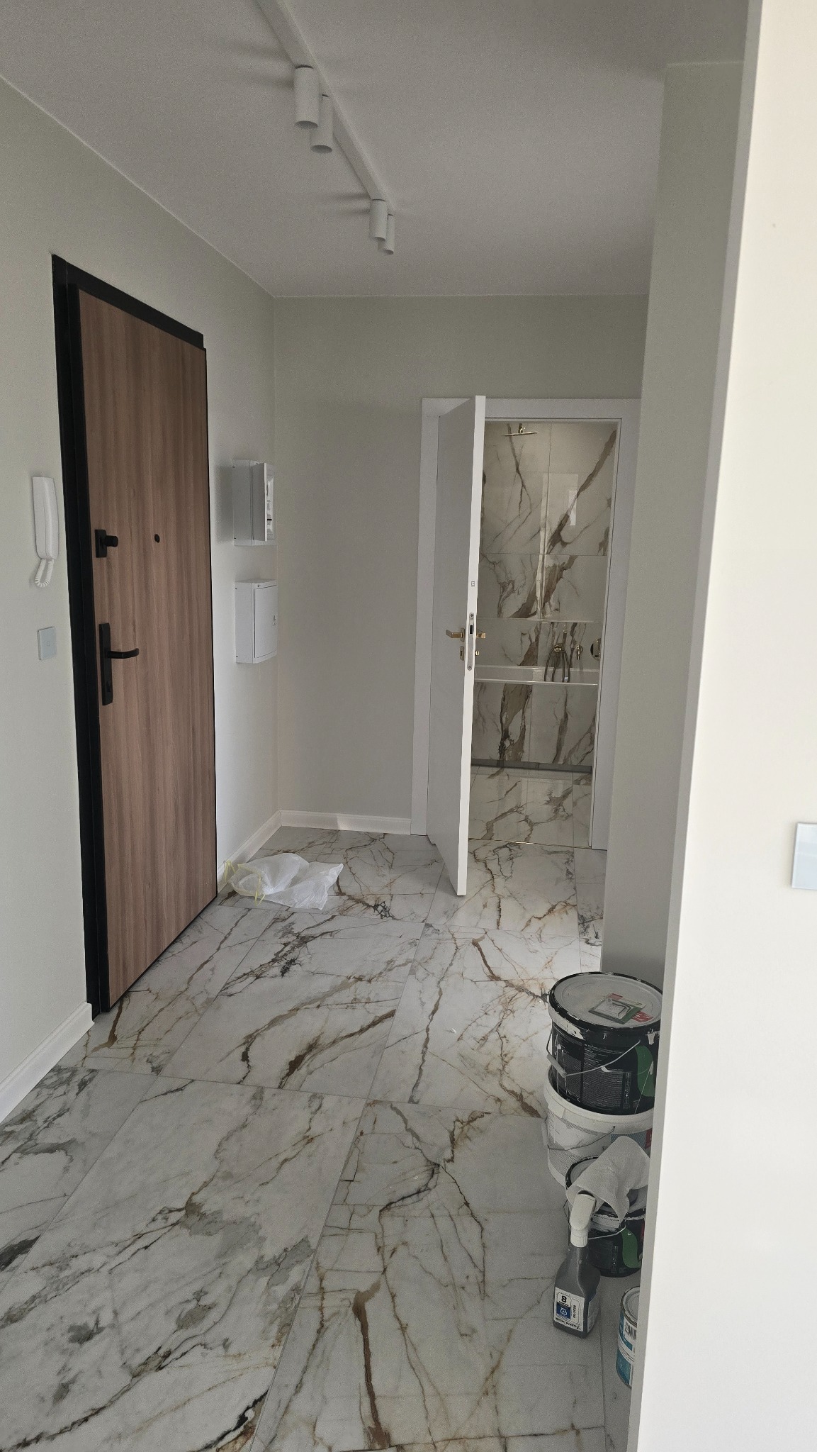 Wnętrze mieszkania w trakcie wykańczania, z widoczną otwartą łazienką z marmurowymi płytkami, nową podłogą imitującą marmur, drewnianymi drzwiami wejściowymi z czarną ramą i wiaderkami z farbą...