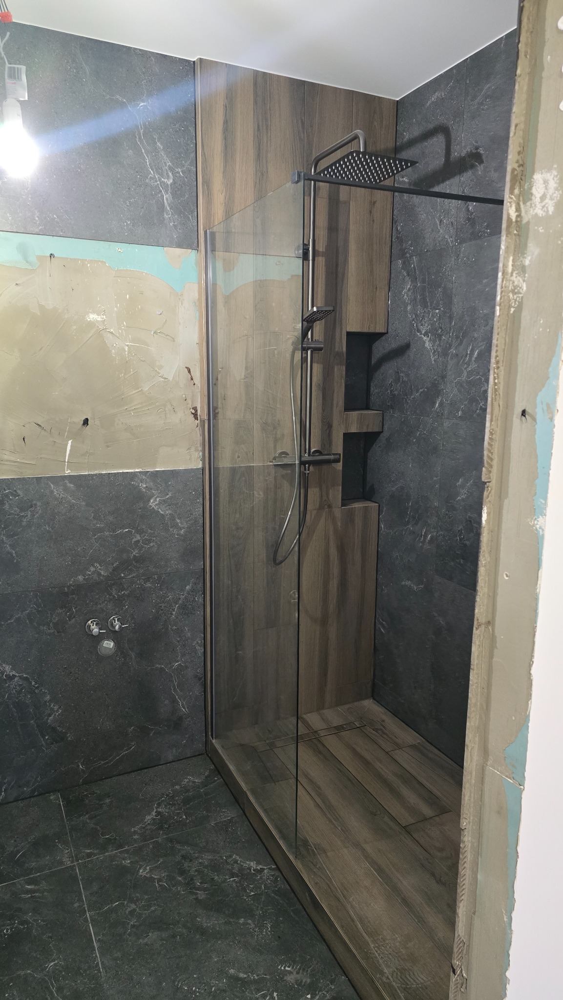 Wnętrze łazienki w trakcie prac: kabina prysznicowa typu walk-in z brązowymi płytkami imitującymi drewno, szara marmurowa okładzina ścienna, widoczne ślady po gładzi na ścianie po lewej stronie.