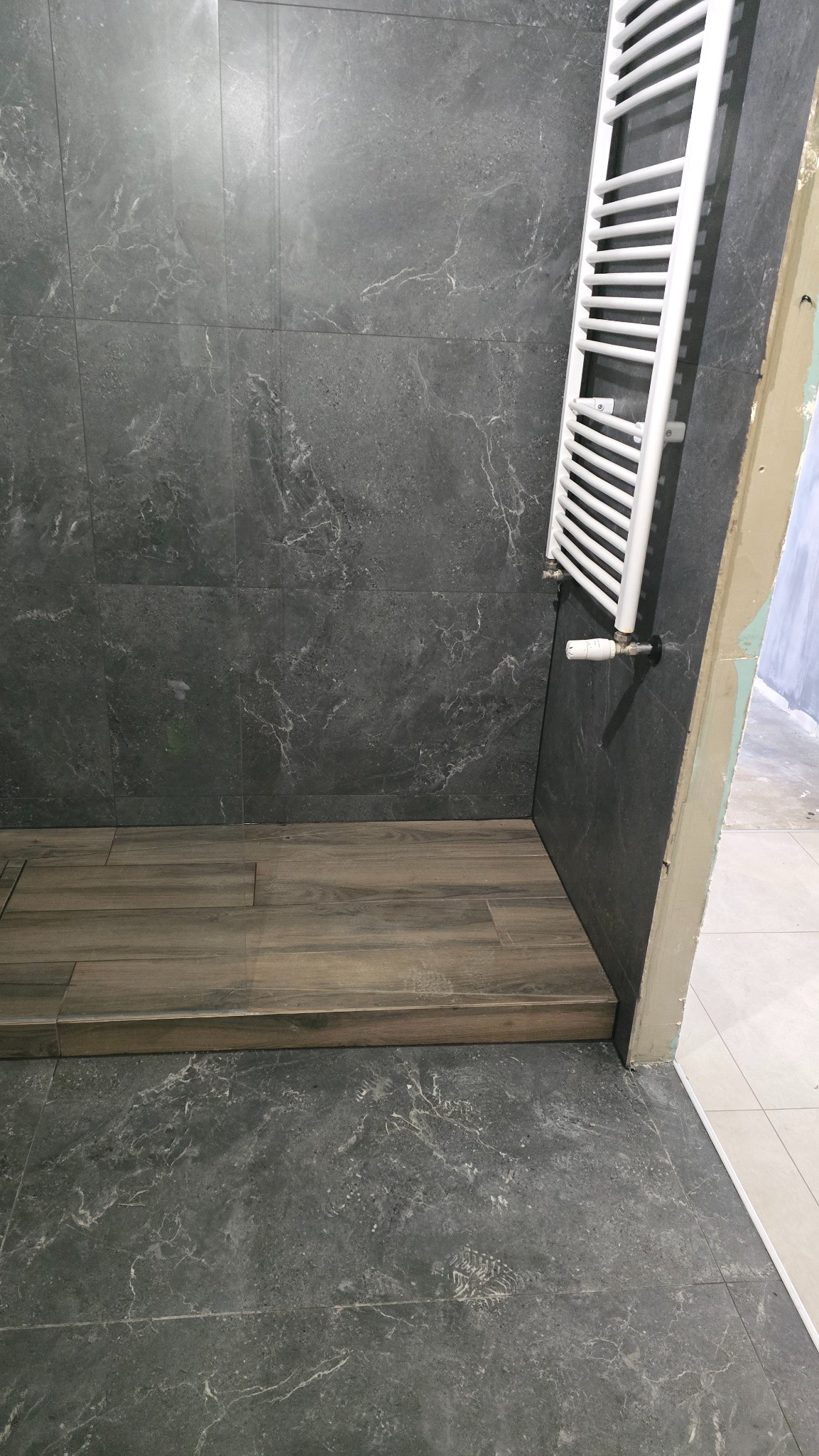 Fragment łazienki w trakcie prac wykończeniowych: ciemnoszare płytki imitujące marmur na ścianach i podłodze, drewnopodobny podest prysznicowy, biały grzejnik drabinkowy zamontowany na ścianie...