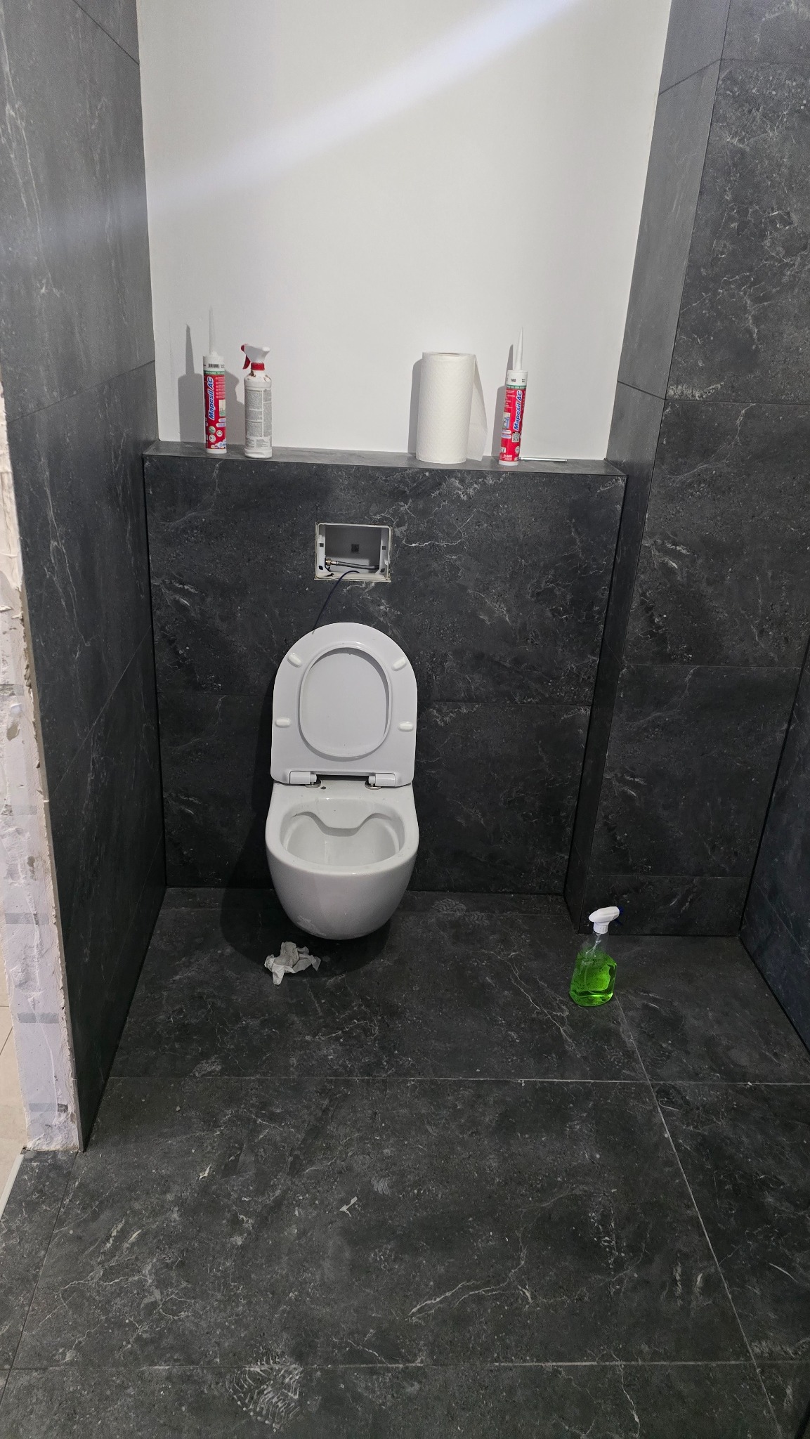 Wnętrze łazienki w trakcie prac w Warszawie: widoczna toaleta z otwartą klapą, czarne płytki imitujące marmur na ścianach i podłodze, oraz akcesoria budowlane i środki czystości na półce. Otwór...