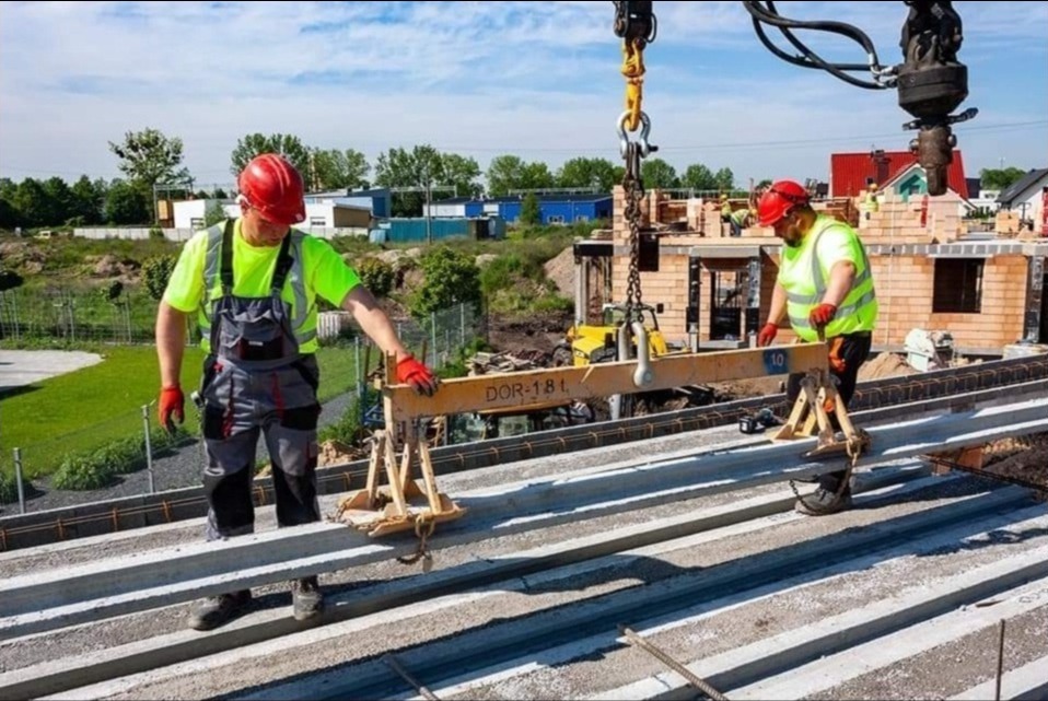 Montaż stropu Teriva: Dwóch pracowników w kaskach i odblaskowych kamizelkach umieszcza prefabrykowane belki stropowe za pomocą dźwigu na budowie domu jednorodzinnego.