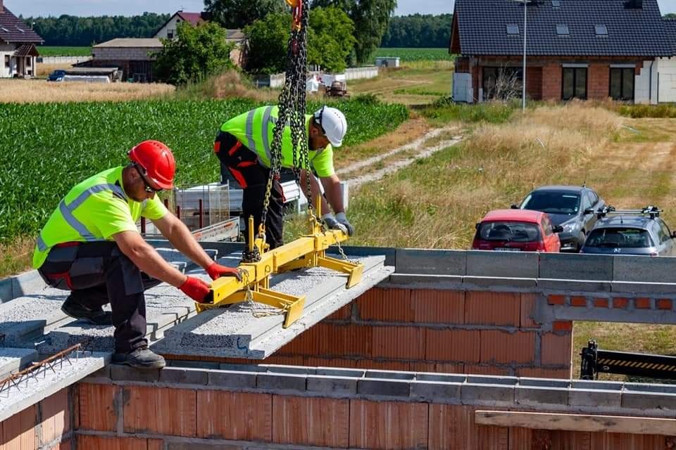 Dwóch pracowników w kamizelkach odblaskowych i kaskach umieszcza betonowy prefabrykat stropowy za pomocą żółtego uchwytu dźwigowego na wznoszone ściany z czerwonej cegły