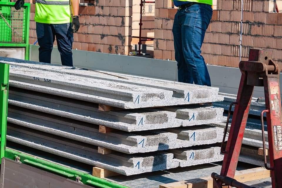 Stos szarych, prefabrykowanych płyt betonowych z oznaczeniami na palecie transportowej, w tle mur z cegły ceramicznej dziurawki i fragmenty sylwetek pracowników w odblaskowych kamizelkach.