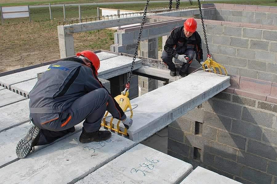 Montaż stropu z płyty strunobetonowej SMART o szerokości 60 cm przez dwóch pracowników w kaskach, z użyciem chwytaka i łańcucha, na budowie z elementami z betonu komórkowego.