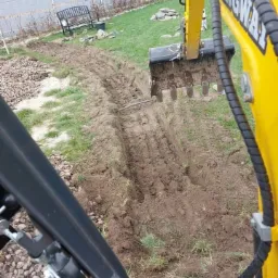 Żółta koparka z łyżką z zębami w trakcie wykonywania wąskiego wykopu w ziemi, widok z kabiny operatora, w tle trawnik i ogrodzenie.