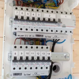 Otwarta, biała skrzynka rozdzielcza z widocznymi bezpiecznikami oznaczonymi numerami i nazwami pomieszczeń (salon dół, łazienka góra, kuchnia), oraz uporządkowanymi przewodami w różnych kolorach...