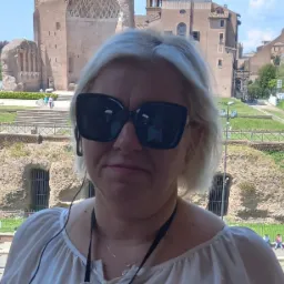 Kobieta w okularach przeciwsłonecznych na tle ruin starożytnego miasta, z widocznymi budowlami z jasnego kamienia i fragmentami zieleni.