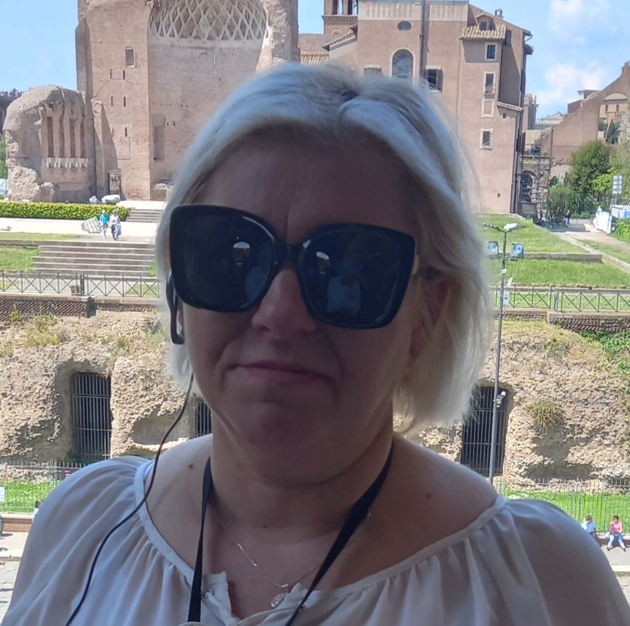 Kobieta w okularach przeciwsłonecznych na tle ruin starożytnego miasta, z widocznymi budowlami z jasnego kamienia i fragmentami zieleni.