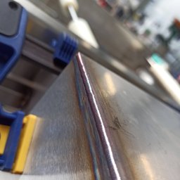 JOGILLA Art Welding - Spoina na metalu, zabezpieczona niebiesko-żółtym ściskiem, wykonana z widocznymi tęczowymi przebarwieniami termicznymi.