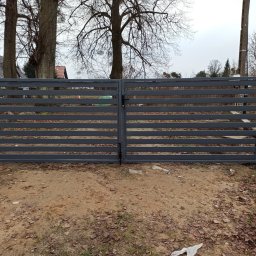 JOGILLA Art Welding - Nowoczesna, dwuskrzydłowa brama wjazdowa z poziomymi profilami, pomalowana na szaro, z widoczną klamką i zamkiem, na tle jesiennego krajobrazu z drzewami bez liści i domem w oddali.