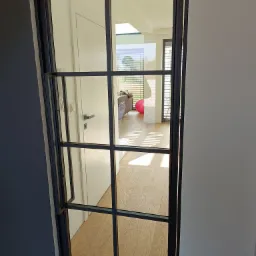 Czarna, przeszklona, metalowa rama drzwi wewnętrznych z podziałami, widok na jasne wnętrze z drewnianą podłogą i elementami dekoracyjnymi.