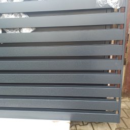 JOGILLA Art Welding - Fragment szarego, metalowego ogrodzenia panelowego z poziomymi lamelami, widoczne elementy opakowania ochronnego i fragment drewnianego ogrodzenia w tle.