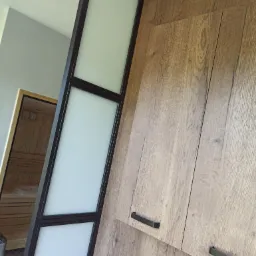 Nowoczesna ścianka działowa z trzema matowymi szybami w czarnej ramie, przylegająca do drewnianej szafki, z widoczną sauną w tle.