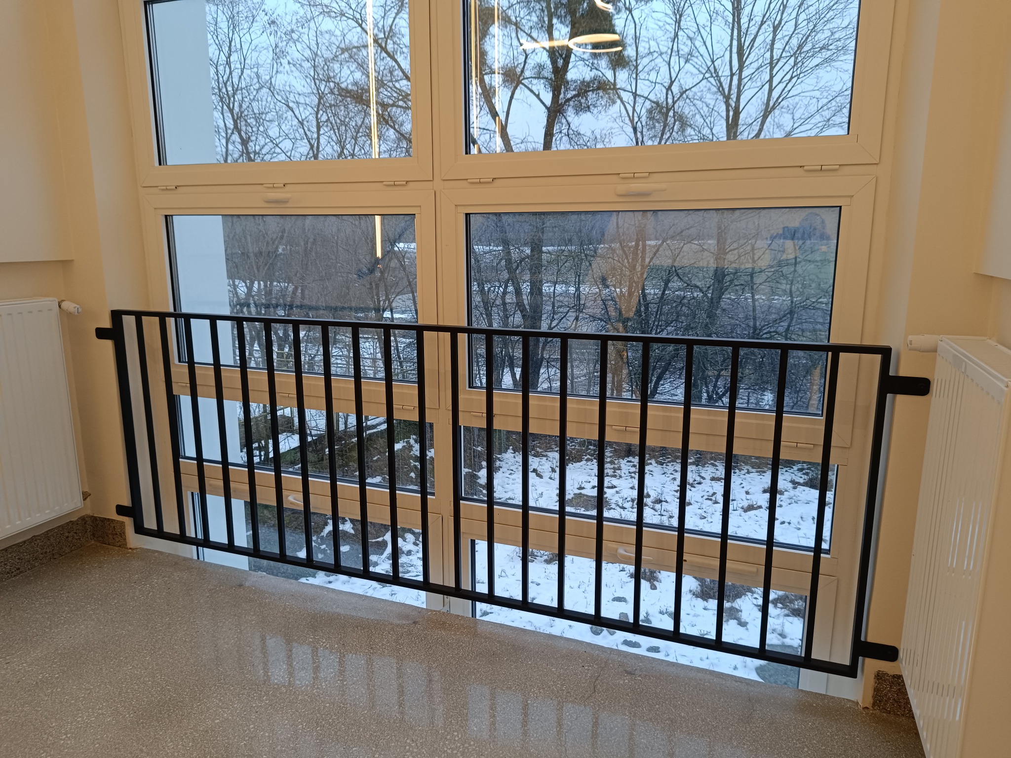 Czarna, metalowa barierka zabezpieczająca okno na podeście, widok na zimowy krajobraz za oknem, grzejnik po prawej stronie kadru.