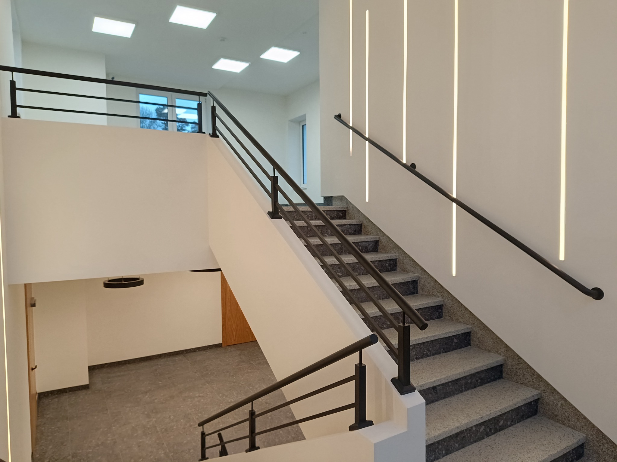 Wnętrze budynku z dwubiegowymi schodami o szarych stopniach z lastryko i czarną, minimalistyczną balustradą z poziomymi prętami. Na ścianie pionowe, dekoracyjne oświetlenie LED.
