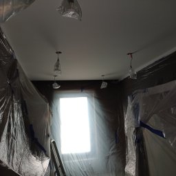 Solidne Malowanie - Przygotowane pomieszczenie do prac wykończeniowych: zabezpieczone folią ściany i wiszące lampy, świeżo pomalowany sufit na biało, drabina oparta o okno.