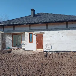 Surowy stan domu jednorodzinnego z białych bloczków, z zamontowanymi oknami i drzwiami wejściowymi, widoczne ślady po oponach na ziemi.