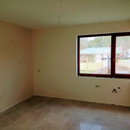 Solidne Malowanie - Pusty pokój po szpachlowaniu i malowaniu, widoczne przygotowane punkty elektryczne i hydrauliczne, świeżo położone płytki podłogowe, okno z widokiem na ogród.