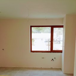 Solidne Malowanie - Wnętrze pokoju po szpachlowaniu i wstępnym malowaniu, widoczne wyprowadzone kable elektryczne i instalacje hydrauliczne pod oknem, ściany w jasnym odcieniu beżu.