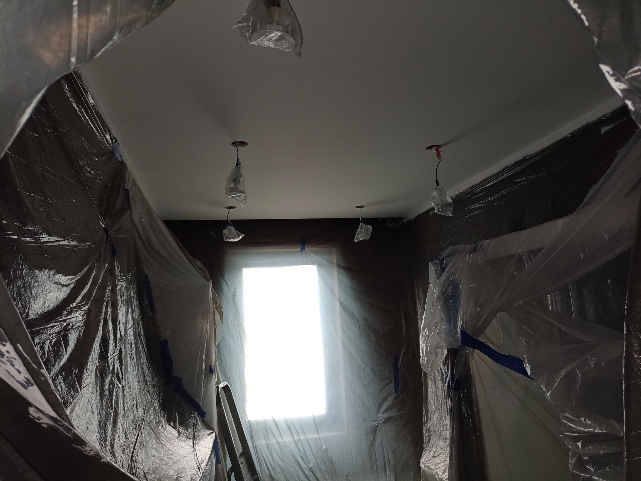 Przygotowane pomieszczenie do prac wykończeniowych: zabezpieczone folią ściany i wiszące lampy, świeżo pomalowany sufit na biało, drabina oparta o okno.