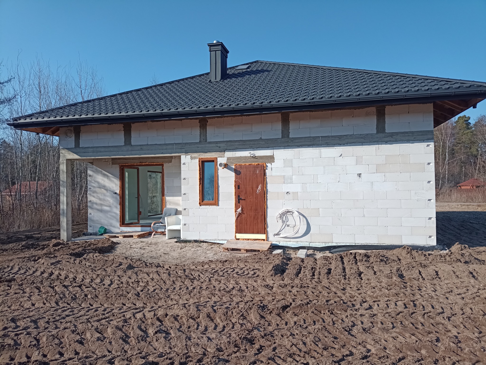 Surowy stan domu jednorodzinnego z białych bloczków, z zamontowanymi oknami i drzwiami wejściowymi, widoczne ślady po oponach na ziemi.