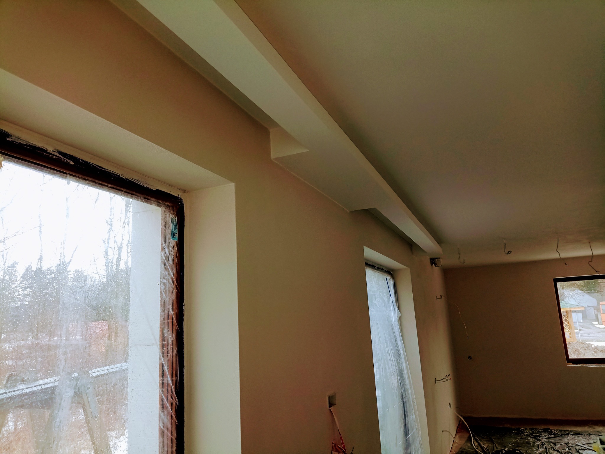 Świeżo wykończone wnętrze z oknami zabezpieczonymi folią malarską, listwy dekoracyjne pod sufitem, przygotowane do dalszych prac wykończeniowych.