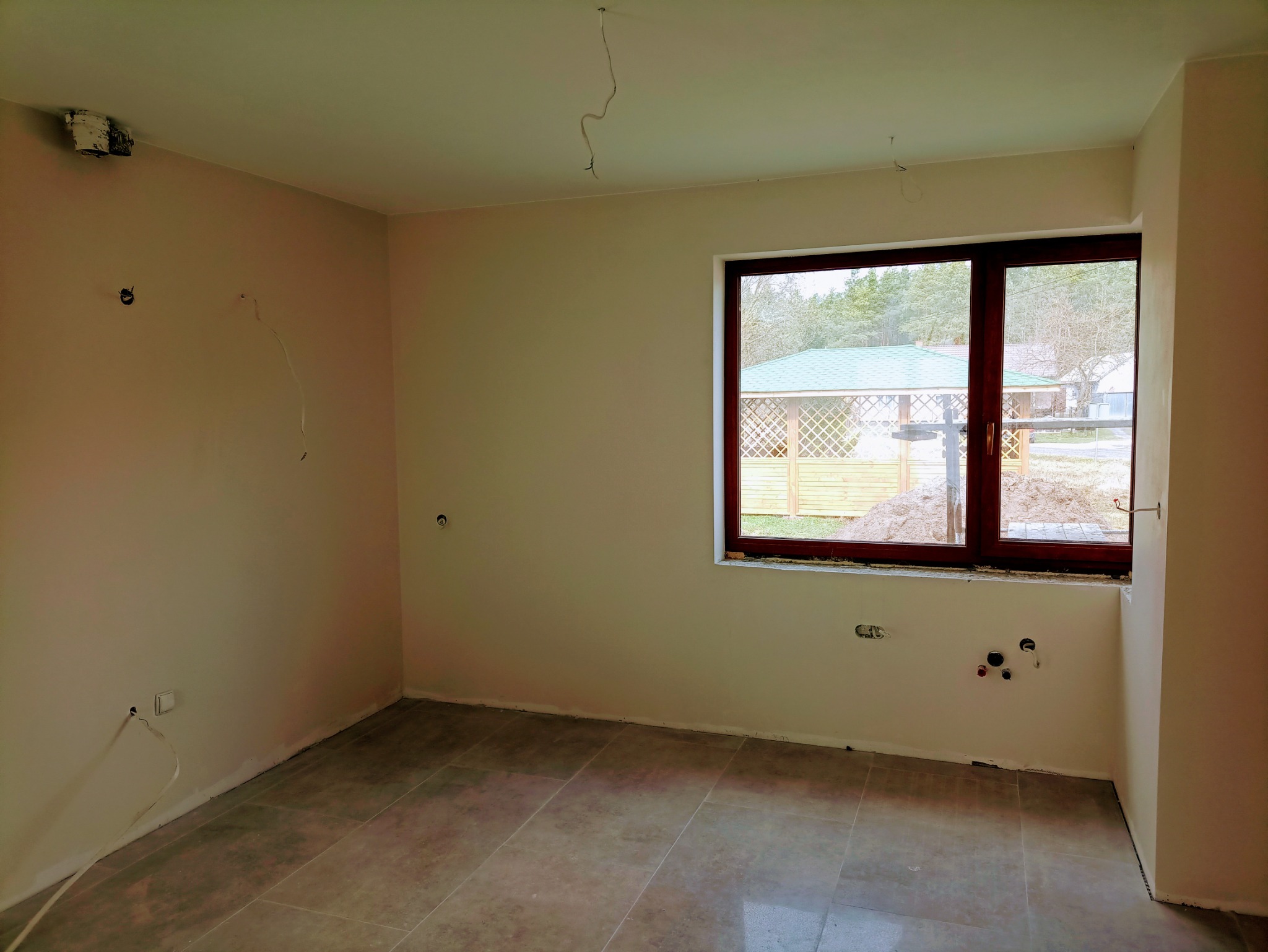 Pusty pokój po szpachlowaniu i malowaniu, widoczne przygotowane punkty elektryczne i hydrauliczne, świeżo położone płytki podłogowe, okno z widokiem na ogród.