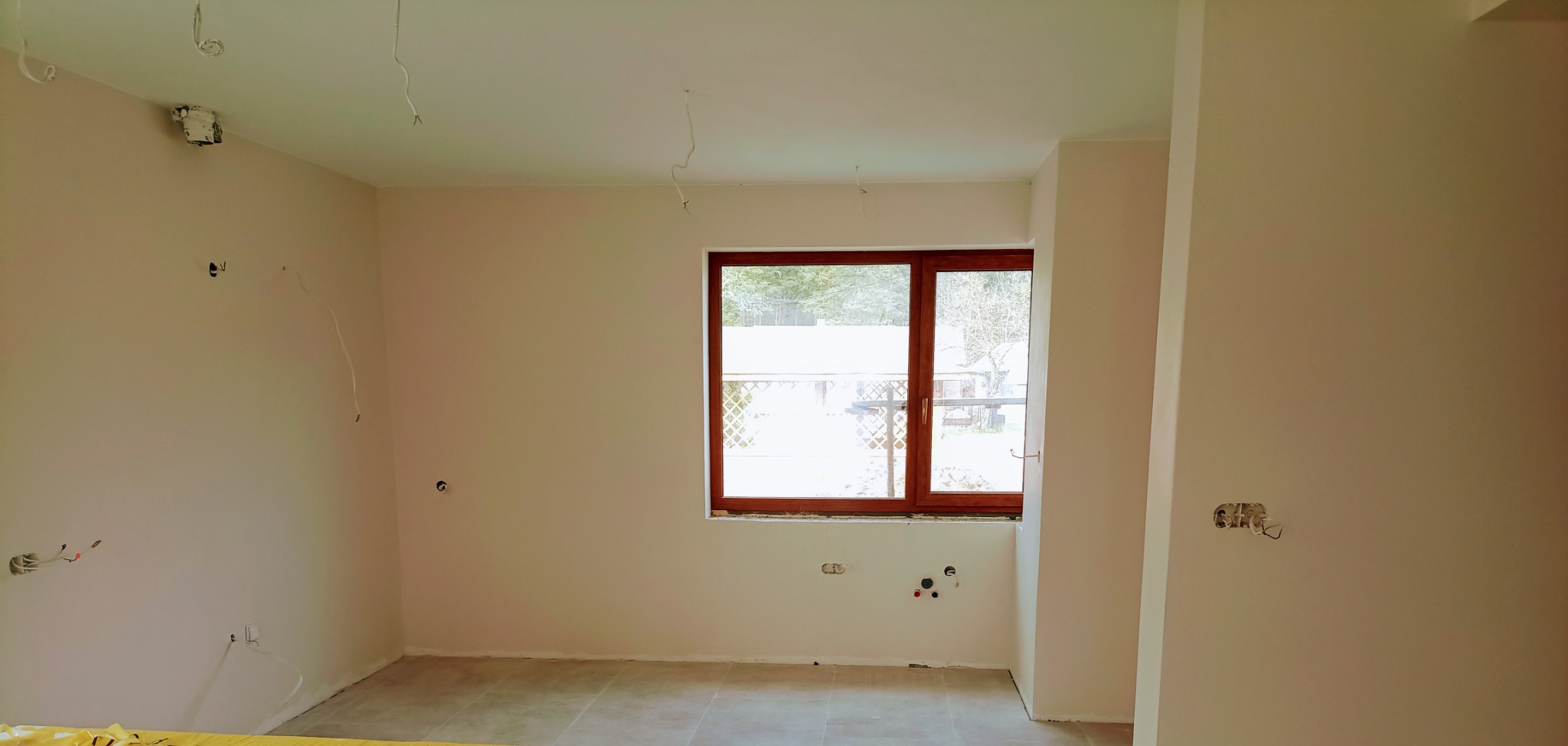Wnętrze pokoju po szpachlowaniu i wstępnym malowaniu, widoczne wyprowadzone kable elektryczne i instalacje hydrauliczne pod oknem, ściany w jasnym odcieniu beżu.