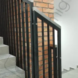 Czarne, pionowe słupki balustrady z nierdzewną górną krawędzią, zamontowane przy ceglanej ścianie klatki schodowej z szarymi stopniami.