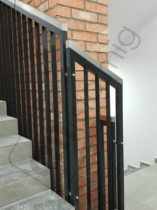 Czarne, pionowe słupki balustrady z nierdzewną górną krawędzią, zamontowane przy ceglanej ścianie klatki schodowej z szarymi stopniami.