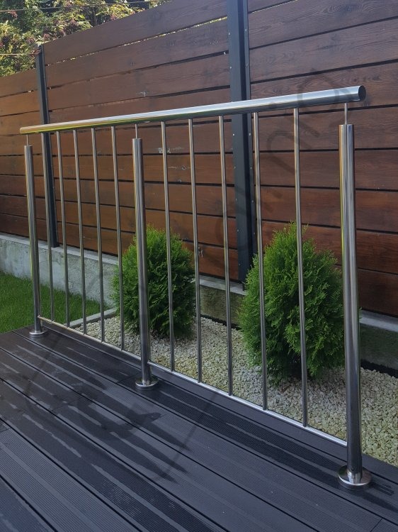 Nierdzewna balustrada ze słupkami na tarasie z desek kompozytowych, widok na drewniany płot i rośliny ozdobne.