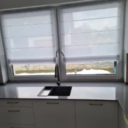 Białe rolety rzymskie na oknach nad szarym blatem kuchennym z czarnym zlewem, widok na ośnieżony ogród.